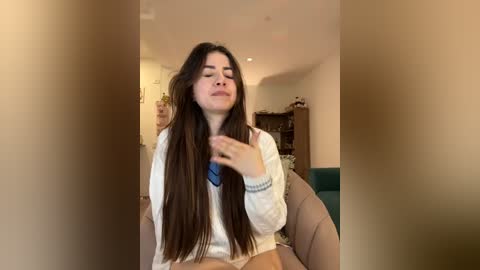 Media: Video of a young woman with long brown hair, wearing a white sweater and a blue necklace, sitting on a teal couch, with a blurred background showing a living room.