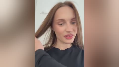 Media: Video of a fair-skinned, young Caucasian woman with straight, light brown hair, smiling, wearing a black top. She's indoors with blurred, neutral-colored background.