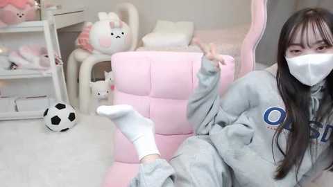 Media: Video of an Asian woman in a light gray hoodie, white mask, and white socks, seated on a pink gaming chair, with a soccer ball on the floor, and plush toys in the background.