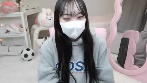 Media: Video of an Asian woman with long black hair, wearing a white mask, grey hoodie, and seated in a pink gaming chair. Background features a white shelf with plush toys, including a white cat, and a pink gaming headset.