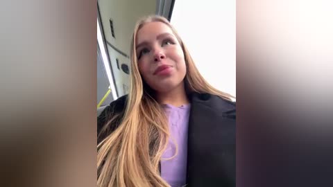 Media: Video of a blonde woman with long hair, wearing a black blazer and light purple shirt, looking out a car window. The background shows the car's interior and outside.