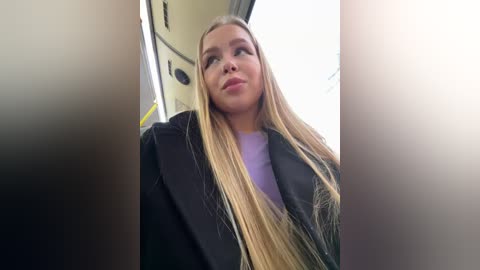 Media: Video of a young Caucasian woman with long, straight blonde hair, wearing a black coat and purple top, gazing upward through a car window, with blurred, sunlit background.