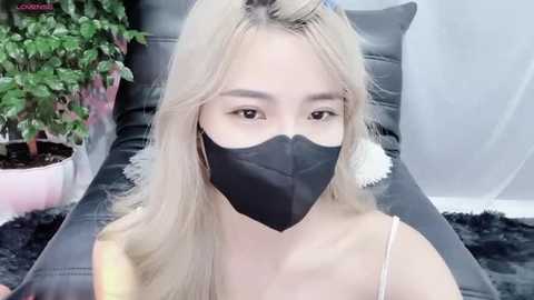 Media: Video of a young woman with long blonde hair, wearing a black face mask and a white camisole, sitting on a black leather couch with a potted plant in the background.