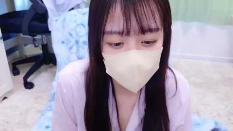 Media: Video of an Asian woman with long black hair and pale skin, wearing a beige surgical mask, kneeling on a carpeted floor in a cluttered office.