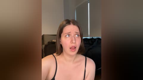 Media: Video of a Caucasian woman with light skin, straight brown hair, and blue eyes, wearing a black spaghetti strap top. She has a surprised expression, mouth open, in a dimly lit room with beige walls and a window.