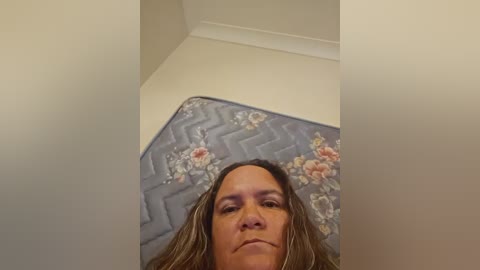 Media: Video of a woman with long brown hair lying on a blue quilted mattress with floral patterns, in a corner of a beige room with white trim.