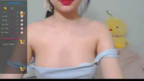 Media: A video of an Asian woman with fair skin, wearing red lipstick, holding her off-shoulder grey top, showing cleavage. Background features a stuffed yellow duck. Live streaming interface overlay on the left.