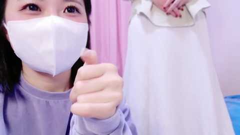 Media: A video of an Asian woman with long black hair and light skin wearing a white surgical mask, giving a thumbs-up gesture. She's in a room with a pink curtain and a white medical gown hanging.