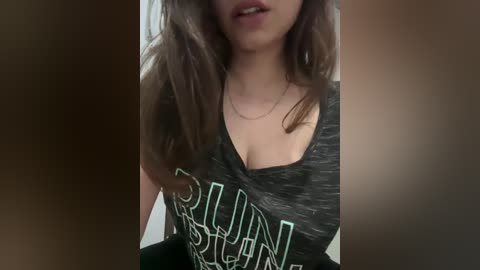 Media: Video of a young woman with long brown hair, wearing a low-cut, gray graphic tee with \"SUN\" in green letters, partially visible cleavage, and a thin gold necklace.