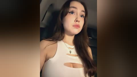 Media: Video of a young Caucasian woman with long brown hair, blue eyes, wearing a white crop top with cutouts, a green necklace, and minimal makeup.