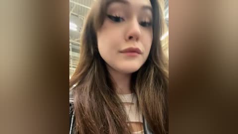 Media: Video of a young woman with fair skin and long, wavy brown hair, wearing a patterned top. She has subtle makeup with dark eyeliner and light lipstick. Background shows a beige wall with a metal ceiling.