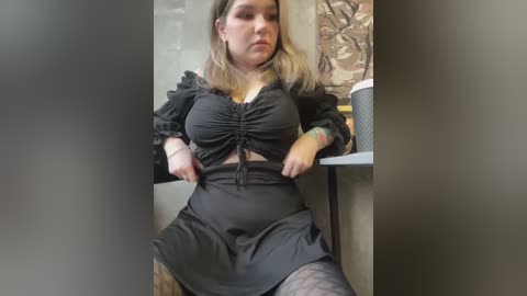 Media: A video of a plus-sized woman with light skin, wearing a black ruffled crop top and black skirt, sitting in a dimly lit room with a wooden wall. She has shoulder-length blonde hair and is holding her skirt.