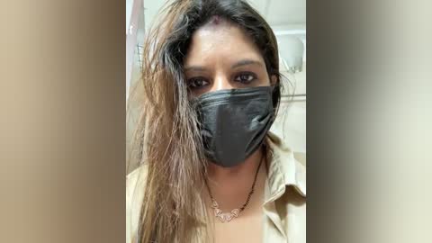 Media: Video of a young South Asian woman with medium-brown skin, wearing a black surgical mask, long brown hair, and a beige shirt. She's indoors, with a blurred background of beige tiles.