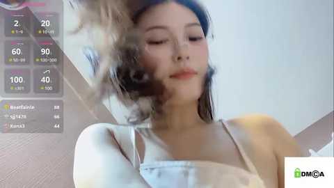 Media: Video of a young Asian woman with long, dark hair, wearing a white tank top, blowing smoke towards the camera. Background shows a weather app with temperature readings.