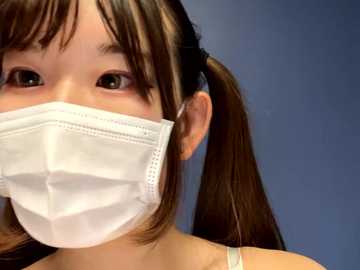 Media: Video of an Asian woman with long, straight brown hair tied in pigtails, wearing a white surgical mask over her nose and mouth. She has light skin and is facing the camera. The background is a solid blue color.