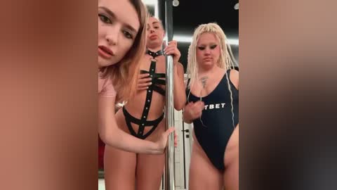 Media: Video of three young women, two in black strappy lingerie, one in a black leotard, standing close together in a dimly lit room with a pole.