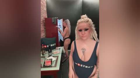 Media: Video of a blonde woman with dreadlocks wearing a black \"Hostess\" one-piece swimsuit, standing in a dimly lit room with a brick wall, a mirror, and a table with Hostess products and a cake.