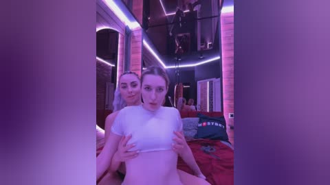 Media: Video of two women in a dimly lit room, one wearing a white crop top, the other a black top, both with light skin and long hair. Background features red and purple neon lights, brick walls, and a bed.