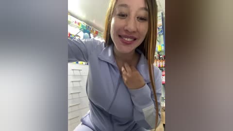 Media: A video of a smiling young woman with long blonde hair, wearing a light blue medical scrub top, standing in a brightly lit pharmacy.