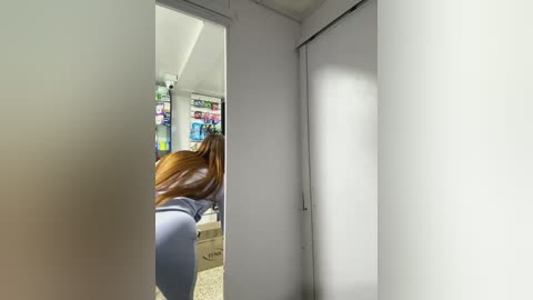 Media: Video of a woman with long brown hair, wearing a light blue top, bending over to pick up something in a bright, modern room with white walls and a carpeted floor.