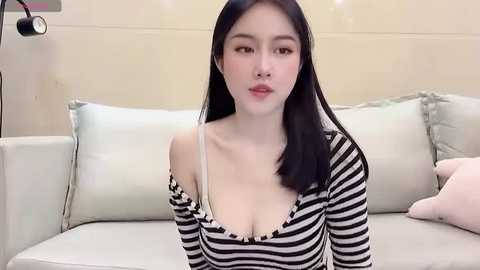 Media: Video of an East Asian woman with straight black hair, fair skin, and light makeup. She wears a low-cut, off-shoulder black and white striped top, sitting on a light-colored couch with pastel pillows.