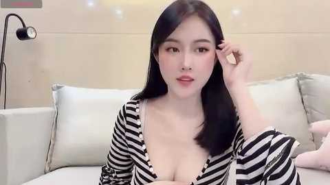 Media: Video of an Asian woman with straight black hair, fair skin, and large breasts in a black and white striped robe, sitting on a beige couch.