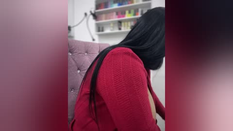 Media: Video of a woman with long black hair and red patterned shirt sitting in a salon chair, with a pink cushioned seat and shelves filled with nail polish bottles in the background.