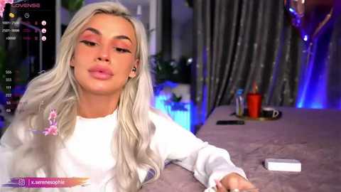 Media: A video of a woman with long, platinum blonde hair, wearing pink eyeshadow and a white long-sleeve shirt, lying on a bed with a gray blanket. The background includes a gray curtain and a red candle.