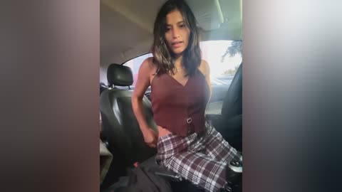 Media: Video of a woman with long dark hair, wearing a sleeveless burgundy top and plaid skirt, sitting in a car, with a blurry background.