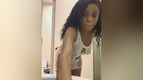 Media: Video of a young Black woman with curly hair, wearing a white crochet tank top, leaning forward in a bathroom mirror selfie.