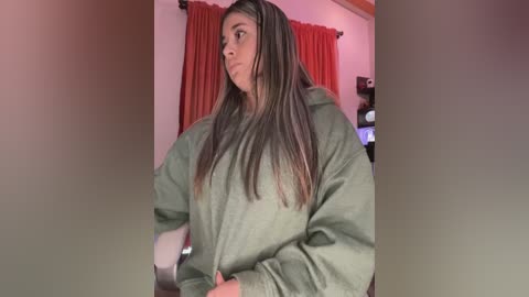 Media: Video of a young woman with long, straight, dark brown hair, wearing a loose-fitting, olive green hoodie, standing indoors with red curtains and shelves in the background.