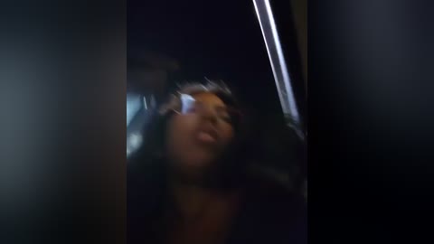 Media: Video of a young Black woman with medium-dark skin, curly hair, and a light complexion. She appears distressed, with her mouth open and eyes closed, possibly in pain or shock. The background is dark and indistinct, suggesting an enclosed space.