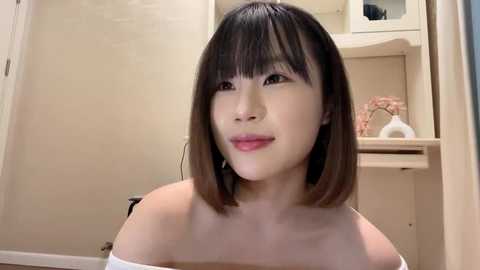 Media: A video of a young Asian woman with straight, shoulder-length black hair and pale skin, wearing a white off-shoulder top, smiling gently. She stands in a beige bathroom with a sink, white towels, and a vase.