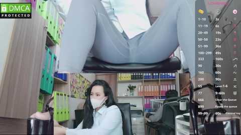 Media: Video of a woman in office attire, wearing a mask, sitting at a desk, with a man's legs in the background, surrounded by colorful folders, indicating a work setting.