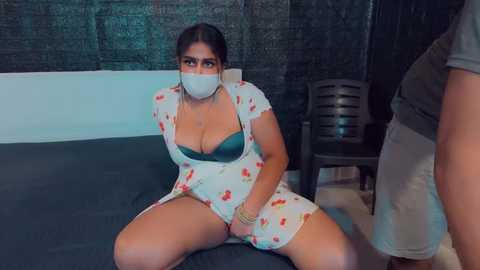 Media: Video of a Latina woman with dark hair and a medium complexion, wearing a white dress with red floral patterns and a green bra, sitting on a bed, gagged, in a dimly lit room.