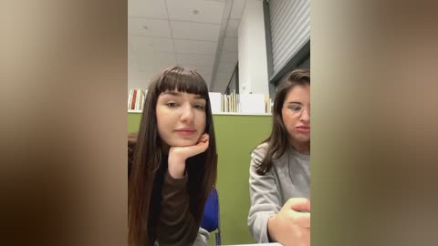 Media: Video of two young women, one with long brown hair and another with dark hair, sitting in a library with a green wall and bookshelves, looking thoughtful.