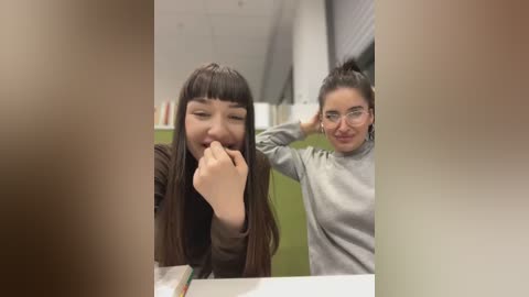 Media: Video of two young women in a modern, brightly lit room. The woman in the foreground, with long brown hair and glasses, is playfully biting her finger. The woman behind her, with a bun and glasses, is smiling.