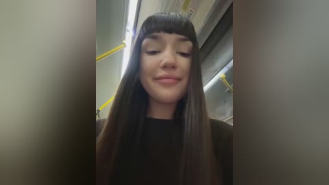 Media: Video of a young woman with long, straight dark hair and blunt bangs, wearing a black top, smiling slightly in an elevator with yellow handrails and gray walls.