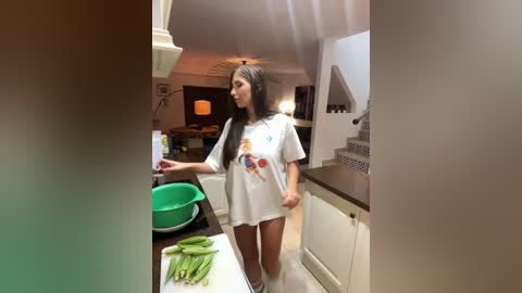 Media: A candid video shows a young woman with long black hair, wearing a white t-shirt and black leggings, standing in a modern kitchen with white cabinets and a green bowl on the counter.