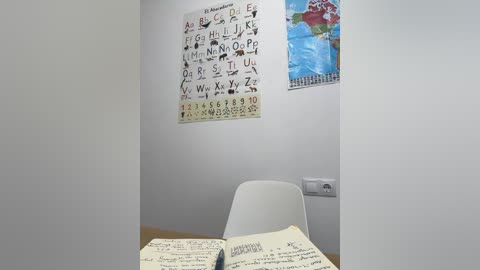 Media: A minimalist classroom photo featuring a white wall with a colorful alphabet chart and a world map, a white chair, and open books on a desk.