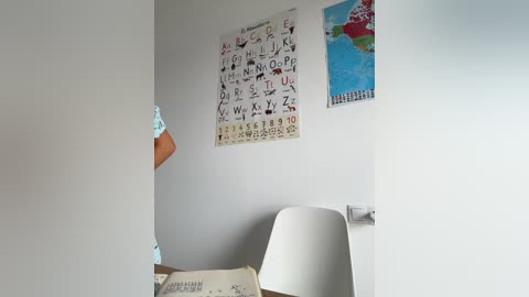 Media: A video of a minimalist, brightly lit room with a white wall displaying a colorful alphabet poster and a world map. A modern white chair and a small, cluttered desk are visible.