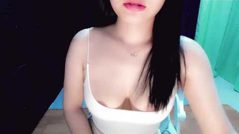 Media: Video of an East Asian woman with long black hair, wearing a white tank top revealing ample cleavage, sitting indoors with a green curtain in the background.