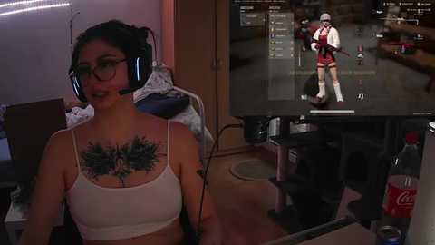 Media: A woman with black hair, glasses, and a white tank top featuring a large flower tattoo on her chest, sits in a gaming chair, wearing a headset. A monitor displays a game with a character in a red and white costume.