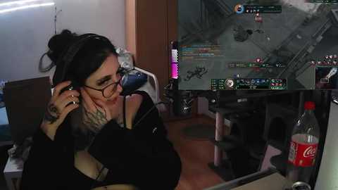 Media: Video of a woman with glasses, black hair in a bun, and tattoos, playing a video game. She is sitting in a dimly lit room with a large TV showing a game screen, and a Coca-Cola bottle on a table.