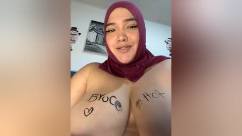 Media: Video of a young, light-skinned woman with a maroon hijab and piercings, smiling. Her bare breasts are marked with tattoos saying \"BRO,\" \"HEART,\" \"HOT,\" and \"HOT.\" A black-and-white picture of a building hangs in the background.