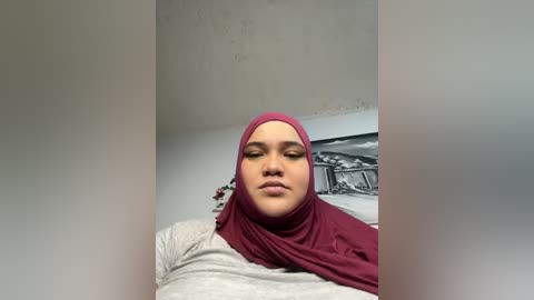 Media: A video of a young woman with light brown skin, wearing a maroon hijab and gray shirt, standing in a dimly lit room with a black-and-white landscape print on the wall.