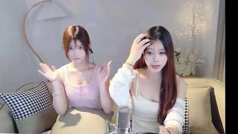 Media: Video of two young Asian women with long hair, one wearing a pink top and the other a white top, sitting on a beige sofa in a dimly lit room.