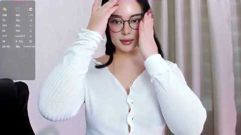 Media: Video of a young woman with fair skin, wearing glasses, adjusting her hair while in a white cardigan, against a beige curtain background.