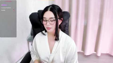 Media: A video of a fair-skinned woman with dark hair, wearing glasses, a white robe, and sitting in a black chair, with a soft pink curtain in the background.