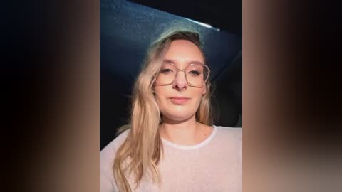 Media: Video of a fair-skinned, blonde-haired woman with glasses, wearing a white, semi-transparent blouse, against a dark, shadowy background.